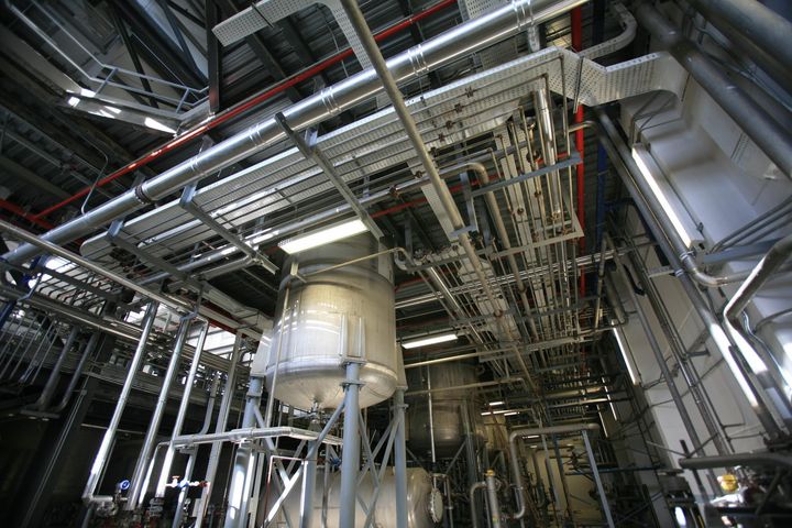 Upward view of closely spaced pipes and cable trays in a production hall, with a cylindrical stainless-steel vessel on supports below.
